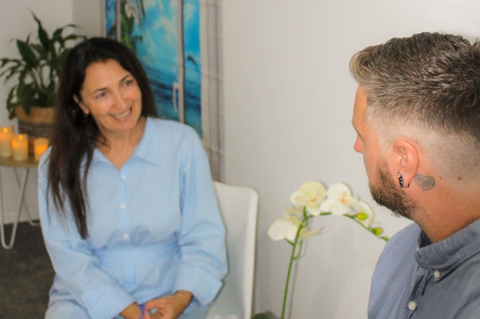 A staff member at Ocean Beach Oxygen in Tauranga, New Zealand, smiling while consulting with a male client. They are in a comfortable, clean therapy room.