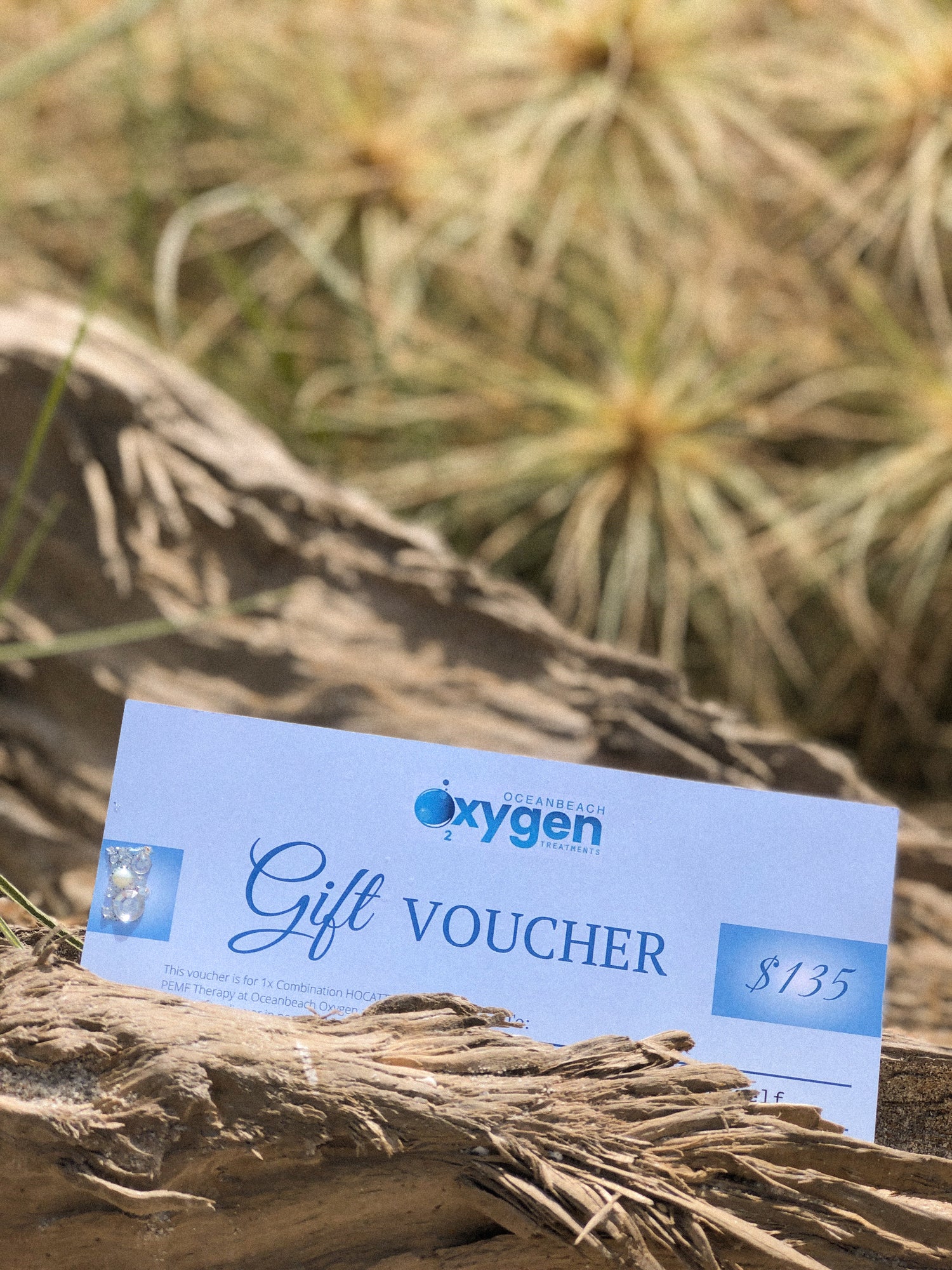 An Oceanbeach Oxygen Treatments gift voucher for $135 rests on a piece of weathered driftwood, suggesting a thoughtful gift for wellness and relaxation.