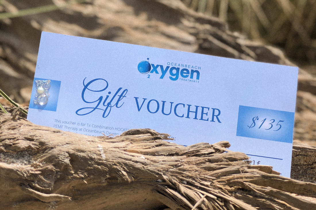 A physical gift voucher from Oceanbeach Oxygen with a value of $135, displayed on a natural background with driftwood and the ocean in the backdrop.